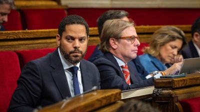 Ignacio Garriga y Joan Garriga de Vox en el Parlament. Ignacio con traje oscuro y corbata azul, Joan con gafas naranjas y traje a rayas. Una mujer con pulsera española mira un dispositivo. Ignacio Garriga y Joan Garriga de Vox en el Parlament. Ignacio con traje oscuro y corbata azul, Joan con gafas naranjas y traje a rayas. Una mujer con pulsera española mira un dispositivo.