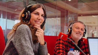 Pastora Vega y Pablo Carbonell, ambos con auriculares, se encuentran en un estudio de radio. Pastora sonríe con un jersey gris, mientras que Pablo viste una camisa de cuadros, frente a un micrófono rojo.