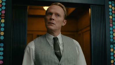 Paul Bettany en una escena de época, con camisa blanca y chaleco gris, mirando sorprendido junto a una entrada con vitrales.