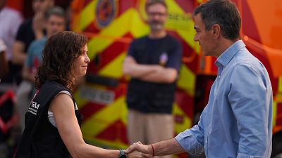 Isabel Díaz Ayuso y Pedro Sánchez se saludan en un evento de emergencias. Ayuso lleva chaleco de "EMERGENCIAS MADRID", Sánchez camisa azul. Vehículo de emergencias al fondo.