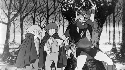 Un fotograma en blanco y negro de una película animada muestra a tres personajes pequeños, posiblemente hobbits, enfrentándose a una figura grande y amenazante en un bosque desolado. La escena sugiere peligro y tensión.