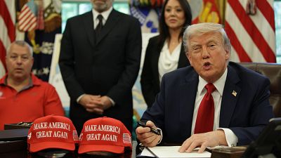 Donald Trump, con traje azul y corbata roja, sentado en un escritorio con gorras "MAKE AMERICA GREAT AGAIN" y banderas al fondo. Donald Trump, con traje azul y corbata roja, sentado en un escritorio con gorras "MAKE AMERICA GREAT AGAIN" y banderas al fondo.