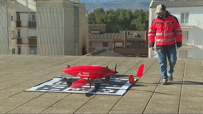 Un dron rojo de la marca "PIRINEOS DRONE" se sitúa sobre una plataforma. Un hombre con ropa de alta visibilidad camina cerca, con edificios y árboles de fondo. Un dron rojo de la marca "PIRINEOS DRONE" se sitúa sobre una plataforma. Un hombre con ropa de alta visibilidad camina cerca, con edificios y árboles de fondo.