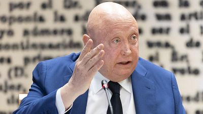 Fernando Samper, con chaqueta azul y corbata negra, gesticula frente a un micrófono. Se observa un texto borroso en el fondo. Fernando Samper, con chaqueta azul y corbata negra, gesticula frente a un micrófono. Se observa un texto borroso en el fondo.