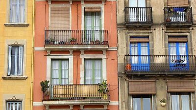 Una fotografía diurna presenta tres edificios contiguos con fachadas de distintos colores: amarillo con contraventanas azules, naranja con ventanas verdes y balcones de hierro, y grisáceo con balcones y contraventanas de madera. Algunos balcones exhiben plantas. Una fotografía diurna presenta tres edificios contiguos con fachadas de distintos colores: amarillo con contraventanas azules, naranja con ventanas verdes y balcones de hierro, y grisáceo con balcones y contraventanas de madera. Algunos balcones exhiben plantas.