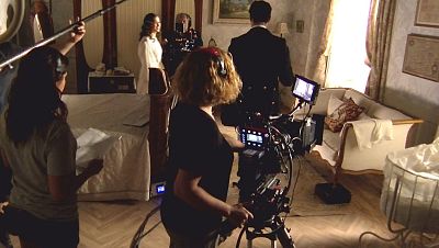 Rodaje de una escena de 'La Promesa': la actriz Carmen Asecas, vestida de época, es grabada por la operadora de cámara y su ayudante de dirección. Se aprecia una iluminación cálida y un decorado de época. Rodaje de una escena de 'La Promesa': la actriz Carmen Asecas, vestida de época, es grabada por la operadora de cámara y su ayudante de dirección. Se aprecia una iluminación cálida y un decorado de época.