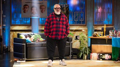 Álex de la Iglesia en un plató de televisión, con camisa roja a cuadros y gafas, de pie ante la cámara.  Detrás, un sofá y una mesa con un premio dorado, fotos y otros objetos.