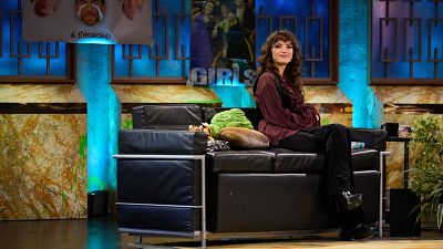 Gabriela Andrada en el plató de "La Revuelta", sentada en un sofá negro con cojines marrones y verdes.  Decorado con paredes de mármol y carteles en la pared.