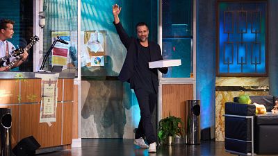 Hugo Silva en el plató de "La Revuelta", saludando y sosteniendo una caja blanca.  Al fondo, un músico toca la guitarra.