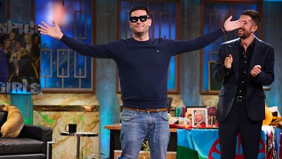 Dos hombres, uno con jersey azul y gafas, el otro con traje, interactúan en un plató de televisión. Uno celebra o saluda, el otro sonríe con complicidad. Dos hombres, uno con jersey azul y gafas, el otro con traje, interactúan en un plató de televisión. Uno celebra o saluda, el otro sonríe con complicidad.