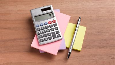 Calculadora plateada encendida mostrando "0", sobre notas adhesivas y bolígrafo plateado, sobre superficie de madera. Sugiere cálculos. Calculadora plateada encendida mostrando "0", sobre notas adhesivas y bolígrafo plateado, sobre superficie de madera. Sugiere cálculos.