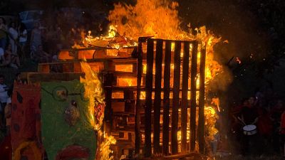 Hoguera nocturna de palés de madera con figuras verdes y rojas consumidas por las llamas, rodeada de espectadores y una persona con tambor. Hoguera nocturna de palés de madera con figuras verdes y rojas consumidas por las llamas, rodeada de espectadores y una persona con tambor.