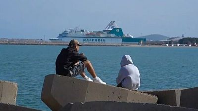 Dos personas sentadas en bloques de hormigón observan el mar. El ferry "DAICADIA" se ve en el horizonte.