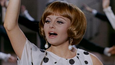 Rocío Dúrcal, con un vestido de lunares y pendientes de perlas, canta con entusiasmo, mientras otras personas vestidas en blanco y negro la acompañan.