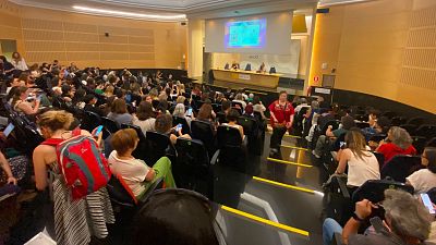 Auditorio moderno repleto de asistentes, mayormente mujeres, observando un escenario con pantalla y el acrónimo "aecid". Panelistas sentadas en una mesa con cartel "AECID".