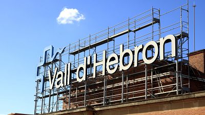 Vista ascendente de un edificio con el letrero "Vall d'Hebron" y andamios metálicos bajo un cielo azul despejado. Vista ascendente de un edificio con el letrero "Vall d'Hebron" y andamios metálicos bajo un cielo azul despejado.