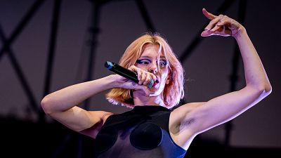 Samantha Hudson, con cabello corto y maquillaje llamativo, canta con un micrófono, vestida con un top negro con detalles. El fondo oscuro difumina el entorno, enfocando la atención en la artista.