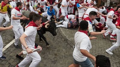 Un toro negro avanza entre corredores vestidos de blanco y rojo en un encierro de San Fermín. La escena muestra el caos y el peligro con personas corriendo y otras en el suelo. Un toro negro avanza entre corredores vestidos de blanco y rojo en un encierro de San Fermín. La escena muestra el caos y el peligro con personas corriendo y otras en el suelo.