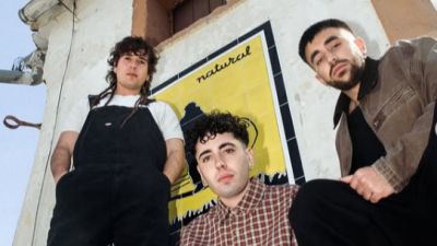 Tres jóvenes se encuentran frente a una pared con azulejos decorativos que muestran la palabra "natural". Uno lleva un mono negro, otro una camisa de cuadros y el tercero una chaqueta marrón.