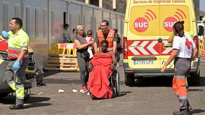 Atención sanitaria a heridos tras el vuelco de un cayuco. Una mujer en silla de ruedas, cubierta con una manta roja, recibe asistencia médica.  Al fondo, una ambulancia.