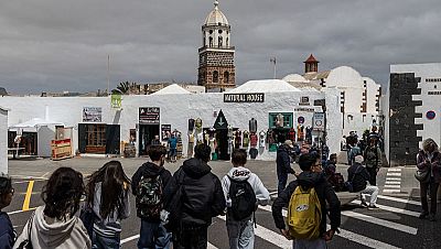 Jóvenes con mochilas caminan por una calle con arquitectura blanca abovedada, posiblemente en Lanzarote. Se observan tiendas y otros turistas. Jóvenes con mochilas caminan por una calle con arquitectura blanca abovedada, posiblemente en Lanzarote. Se observan tiendas y otros turistas.