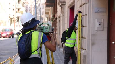 Técnicos realizan mediciones topográficas en una calle urbana, uno con estación total y otro con mira, frente a un edificio con cartel "Luxor". Técnicos realizan mediciones topográficas en una calle urbana, uno con estación total y otro con mira, frente a un edificio con cartel "Luxor".