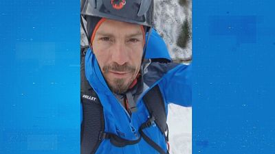 Un hombre con barba y bigote, equipado con casco y ropa de montaña, posa frente a un paisaje nevado. El deportista lleva un casco negro con el logo de una marca y una chaqueta azul. Un hombre con barba y bigote, equipado con casco y ropa de montaña, posa frente a un paisaje nevado. El deportista lleva un casco negro con el logo de una marca y una chaqueta azul.