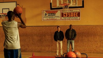 Tres hombres en una cancha de baloncesto interior. Uno lanza, dos observan. Carteles en francés anuncian servicios de mecánica y construcción. Fotograma de 'El sueño americano'. Tres hombres en una cancha de baloncesto interior. Uno lanza, dos observan. Carteles en francés anuncian servicios de mecánica y construcción. Fotograma de 'El sueño americano'.