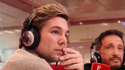 Carlos Cuevas, con el pelo rubio peinado hacia atrás y auriculares, se encuentra en un estudio de radio. Un micrófono rojo con el logo de RNE es visible, junto a otra persona con auriculares.