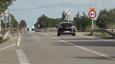Un coche oscuro con matrícula 5509 JGC circula por una carretera de doble sentido, donde una señal limita la velocidad a 80 km/h. El entorno muestra vegetación y un cielo nublado, con la carretera aparentemente mojada.