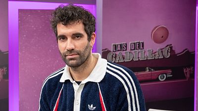 Tamar Novas, con barba y pendiente, posa con una chaqueta deportiva azul marino con detalles blancos y rojos. El fondo morado muestra el nombre de un videopodcast y la silueta de un coche.