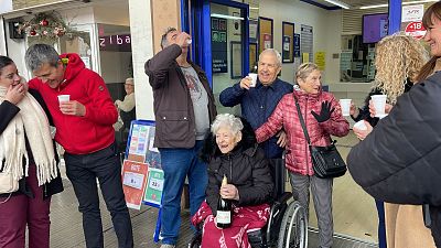 Celebración festiva tras un premio de lotería. Brindis, alegría y una mujer en silla de ruedas con una botella de cava. Cartel con números 8 y 22. Celebración festiva tras un premio de lotería. Brindis, alegría y una mujer en silla de ruedas con una botella de cava. Cartel con números 8 y 22.