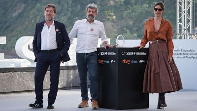 Tres personas posan para una foto en un evento al aire libre. Un hombre con traje azul, otro con camisa y vaqueros, y una mujer con gafas de sol y falda marrón, se apoyan en un atril con logos, con un paisaje montañoso y una estructura blanca de fondo.