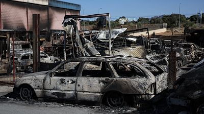 Vehículos calcinados y estructuras dañadas por un incendio, con un cartel "HOLER" al fondo, bajo un cielo azul. Vehículos calcinados y estructuras dañadas por un incendio, con un cartel "HOLER" al fondo, bajo un cielo azul.