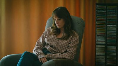 Actriz sentada en sillón gris, camisa a cuadros, vaqueros oscuros, expresión seria. Ambiente íntimo y melancólico.