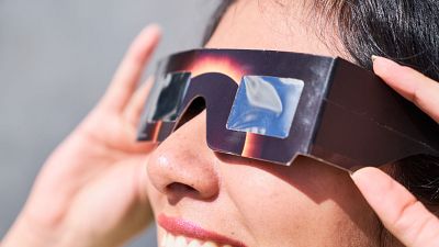 Primer pla del perfil d'una dona amb pèl fosc i maquillatge rosa, ajustant unes ulleres d'eclipsi solar de cartró amb filtres foscos. El fons és gris clar desenfocat. Primer pla del perfil d'una dona amb pèl fosc i maquillatge rosa, ajustant unes ulleres d'eclipsi solar de cartró amb filtres foscos. El fons és gris clar desenfocat.