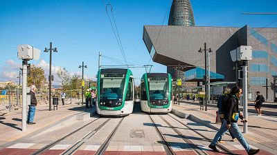 Dos tranvías verdes y blancos con la palabra "TRAM" en el frontal están detenidos en la estación de Glòries, Barcelona, bajo un cielo azul. Personas y la Torre Glòries completan la escena urbana. Dos tranvías verdes y blancos con la palabra "TRAM" en el frontal están detenidos en la estación de Glòries, Barcelona, bajo un cielo azul. Personas y la Torre Glòries completan la escena urbana.