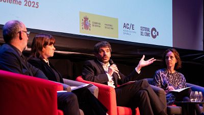 Presentación sobre cómic español: cuatro personas en una mesa redonda, un ponente habla con micrófono. Se ven logotipos del Gobierno de España y el año 2025. Presentación sobre cómic español: cuatro personas en una mesa redonda, un ponente habla con micrófono. Se ven logotipos del Gobierno de España y el año 2025.
