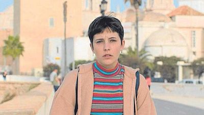 Una joven con cabello oscuro y camiseta de rayas, posiblemente en un fotograma de una película, posa en un entorno exterior con edificios y palmeras, llevando una mochila.