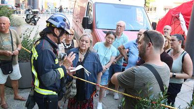Bombero informa a civiles en calle urbana con vegetación, furgoneta y carpa roja. Cinta de balizamiento indica zona restringida. Bombero informa a civiles en calle urbana con vegetación, furgoneta y carpa roja. Cinta de balizamiento indica zona restringida.