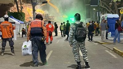 Noche de rescate en Caracas: Protección Civil Chacao, personal con chaleco naranja y militar trabajan entre escombros y excavadoras amarillas, iluminados por luces verdes. Noche de rescate en Caracas: Protección Civil Chacao, personal con chaleco naranja y militar trabajan entre escombros y excavadoras amarillas, iluminados por luces verdes.