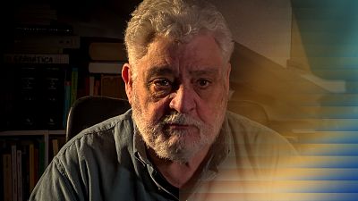 Un hombre mayor, Vicente Romero, con barba blanca y camisa clara, posa frente a una estantería llena de libros, uno de ellos con la palabra "Gramática". La luz, proveniente de una ventana con persianas, crea un juego de luces y sombras. Un hombre mayor, Vicente Romero, con barba blanca y camisa clara, posa frente a una estantería llena de libros, uno de ellos con la palabra "Gramática". La luz, proveniente de una ventana con persianas, crea un juego de luces y sombras.