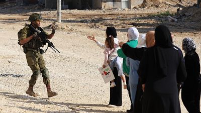 Un soldado israelí impide el paso a un grupo de mujeres y niños. Una niña porta un cartel con símbolos y escritura. El fondo muestra un terreno desolado y construcciones.