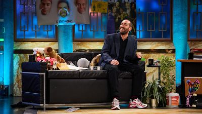 Humorista sentado en un sofá, sonriendo, con chaqueta oscura y pantalones negros. En el set hay peluches, una taza y una planta. Humorista sentado en un sofá, sonriendo, con chaqueta oscura y pantalones negros. En el set hay peluches, una taza y una planta.