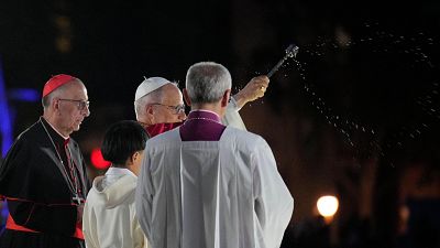 El Papa Francisco, de perfil, esparce agua bendita con un hisopo. A su lado, un cardenal con solideo rojo y un clérigo de espaldas con estola morada completan la escena religiosa. El Papa Francisco, de perfil, esparce agua bendita con un hisopo. A su lado, un cardenal con solideo rojo y un clérigo de espaldas con estola morada completan la escena religiosa.