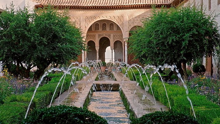 Jardín histórico con canal de agua, fuentes, setos, árboles y edificación islámica con arcos decorados.
