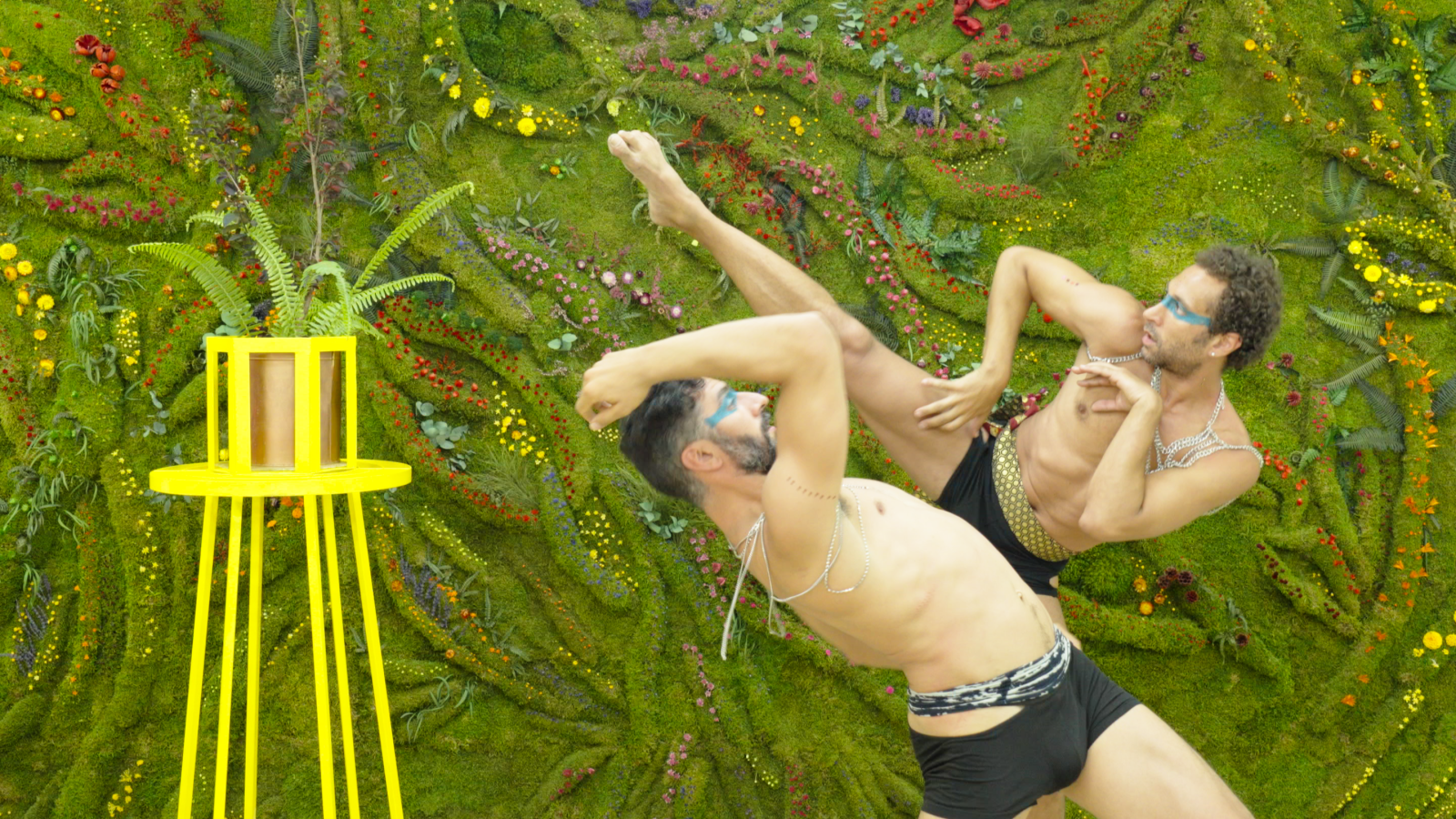Dos bailarines en el Laboratorio Escénico del Bionic Festival de Honevo realizan una coreografía con pintura facial y cadenas plateadas frente a un muro vegetal.