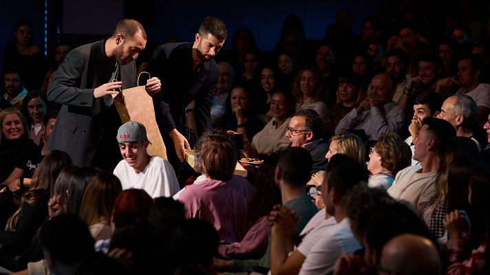 David Broncano y otro hombre entregan una bolsa y comida a un espectador con gorra en un teatro lleno de público.  Escena de un programa de televisión.