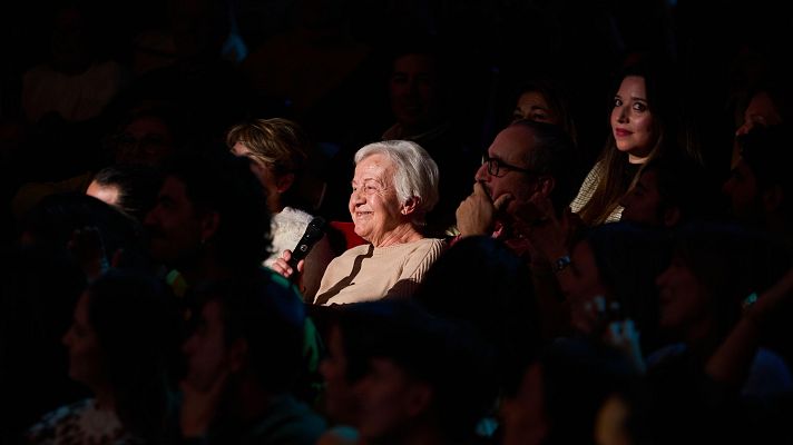 Mujer de cabello canoso sonriendo en un evento festivo, posiblemente su cumpleaños número 90, en un auditorio o teatro.