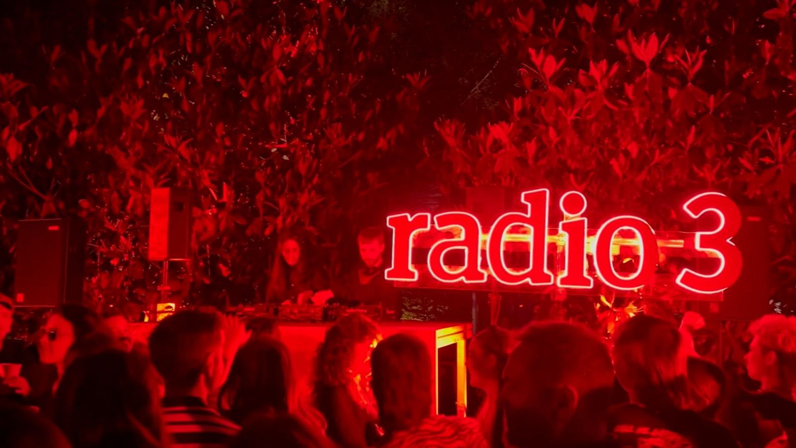 Evento nocturno iluminado en rojo.  Letrero "Radio 3", dos personas tras una mesa de mezclas y público.  Ambiente festivo.